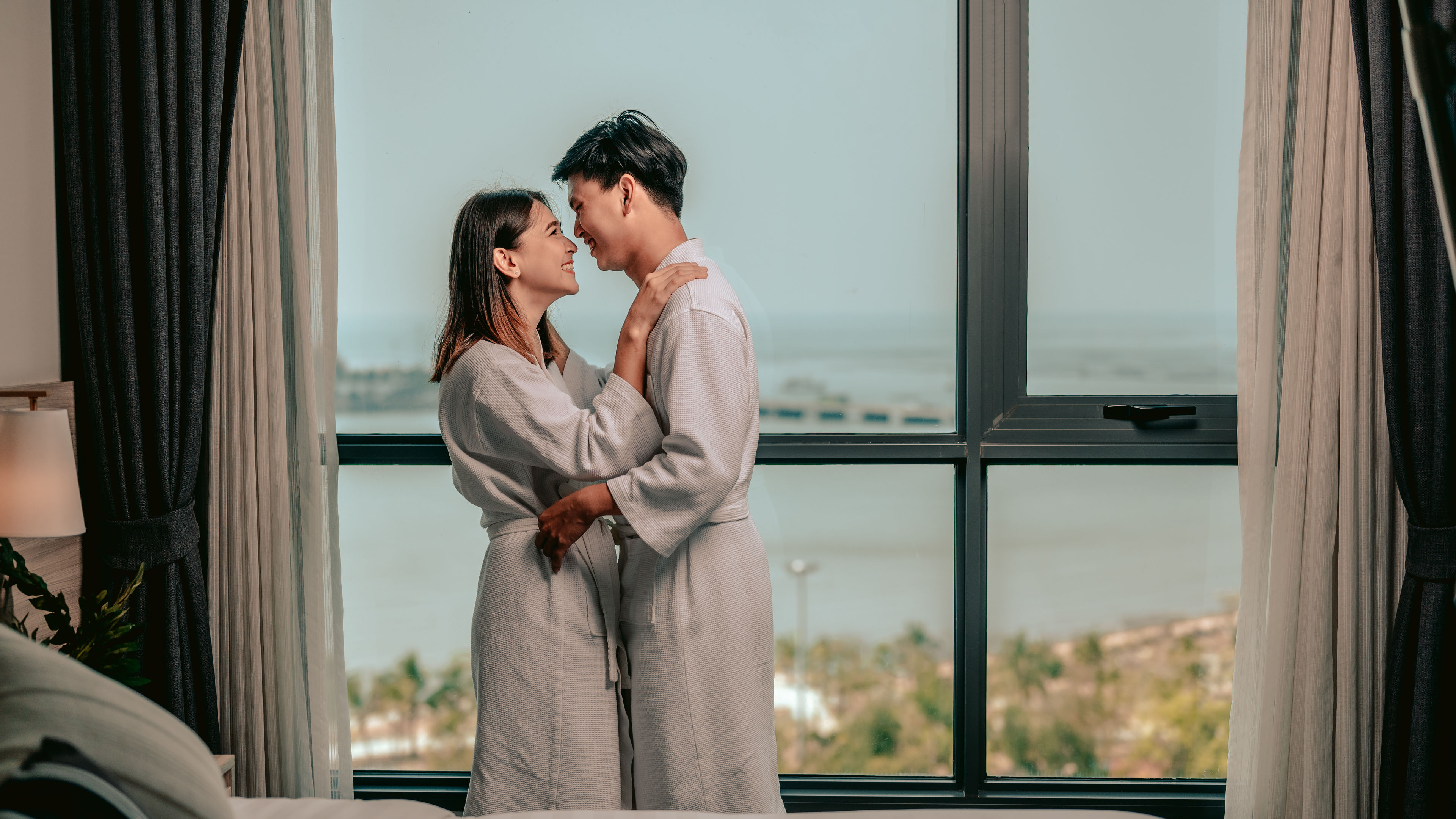 Asian couple is kissing their cheeks in window in their bedroom.